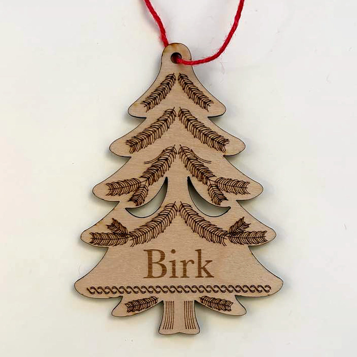 Evergreen tree Christmas ornament in laser-cut birch wood, engraved with the sample name Birk, hanging from a red cord